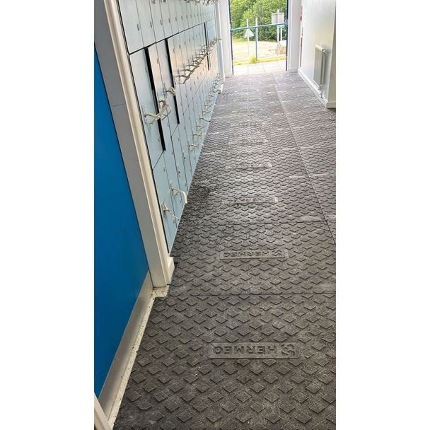 Black plastic ground mats with a raised diamond tread pattern and HERMEQ branding, installed in a hallway beside lockers.