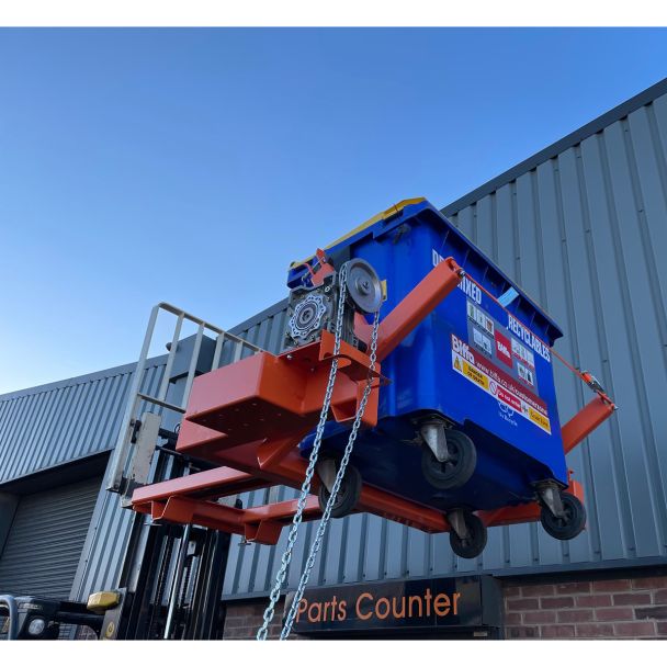 Raised forklift attachment with industrial wheelie bin tipper lifting large recycling bin for waste management operations.